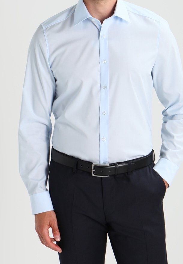 Homme portant une chemise bleu clair à manches longues avec boutons, ceinture noire et pantalon foncé, une main dans la poche.