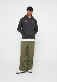 Felpa con cappuccio grigia e logo rosso, abbinata a pantaloni larghi verde oliva. Il modello indossa un cappellino nero e sneaker bianche.
