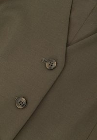 Primo piano del risvolto di un blazer marrone con due bottoni in tartaruga con la scritta "REISS" e dettagli di cucitura visibili.