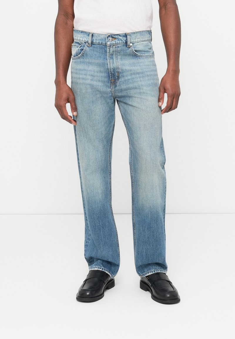 Jasnoniebieskie dżinsy o prostych nogawkach z przetarciami, pięcioma kieszeniami i metalowym zapięciem na guzik. Noszone z czarnymi butami.