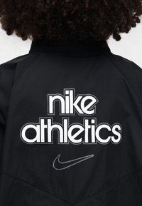 Veste de sport noire avec un grand logo blanc "nike athletics" au dos, et un petit Swoosh Nike gris près de l'ourlet.