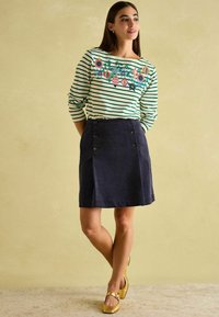 Pruhovaná zeleno-bílá dlouhá košile s barevným vyšíváním. Námořnická kordová sukně s knoflíkovými akcenty a předním rozparkem. Zlaté baleríny.