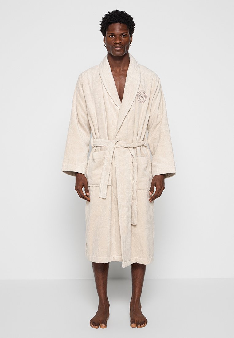 Mężczyzna stoi boso, ubrany w beżowy szlafrok do kolan z związanym paskiem, dwoma przednimi kieszeniami i haftowanym logo na piersi.