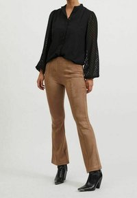 Blouse noire texturée avec devant boutonné et manches bouffantes transparentes, associée à un pantalon ajusté brun effet daim et des bottines noires à talons.
