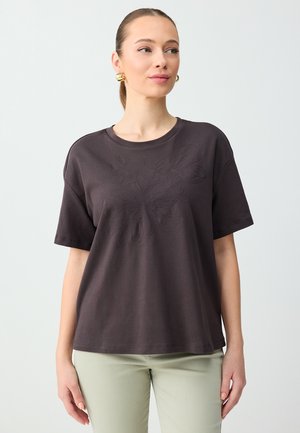 Kobieta ubrana w ciemnobrązową koszulkę z krótkim rękawem z delikatnym kwiatowym haftem, jasne spodnie, złote kolczyki koła, stojąca na jednolitym tle.