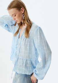 Blusa button-up azzurro chiaro in tessuto a quadri trasparente, maniche lunghe e vita a peplo, con dettaglio a coulisse e una texture sottile.