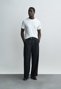 Mann steht mit den Händen in den Taschen, trägt ein weißes Kurzarmhemd, schwarze plissierte Hose, schwarzen Gürtel und schwarze Schuhe vor einem einfarbigen Hintergrund.