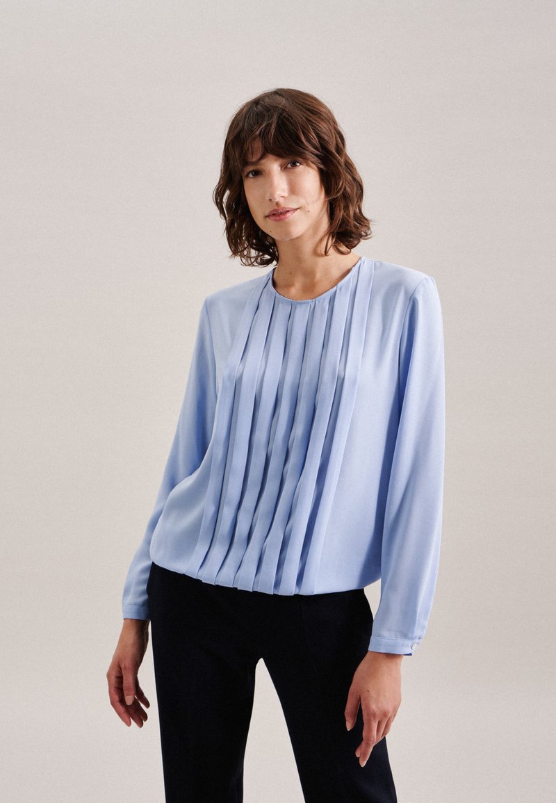 Blusa azzurra chiara con maniche lunghe e pieghe verticali sulla parte frontale, realizzata in tessuto morbido. Collo rotondo con una leggera lucentezza.