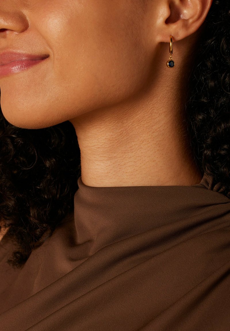 Gouden creool met een enkele blauwe edelsteenhanger. De oorbel heeft een gladde, metalen afwerking en hangt dichtbij het oor van het model.