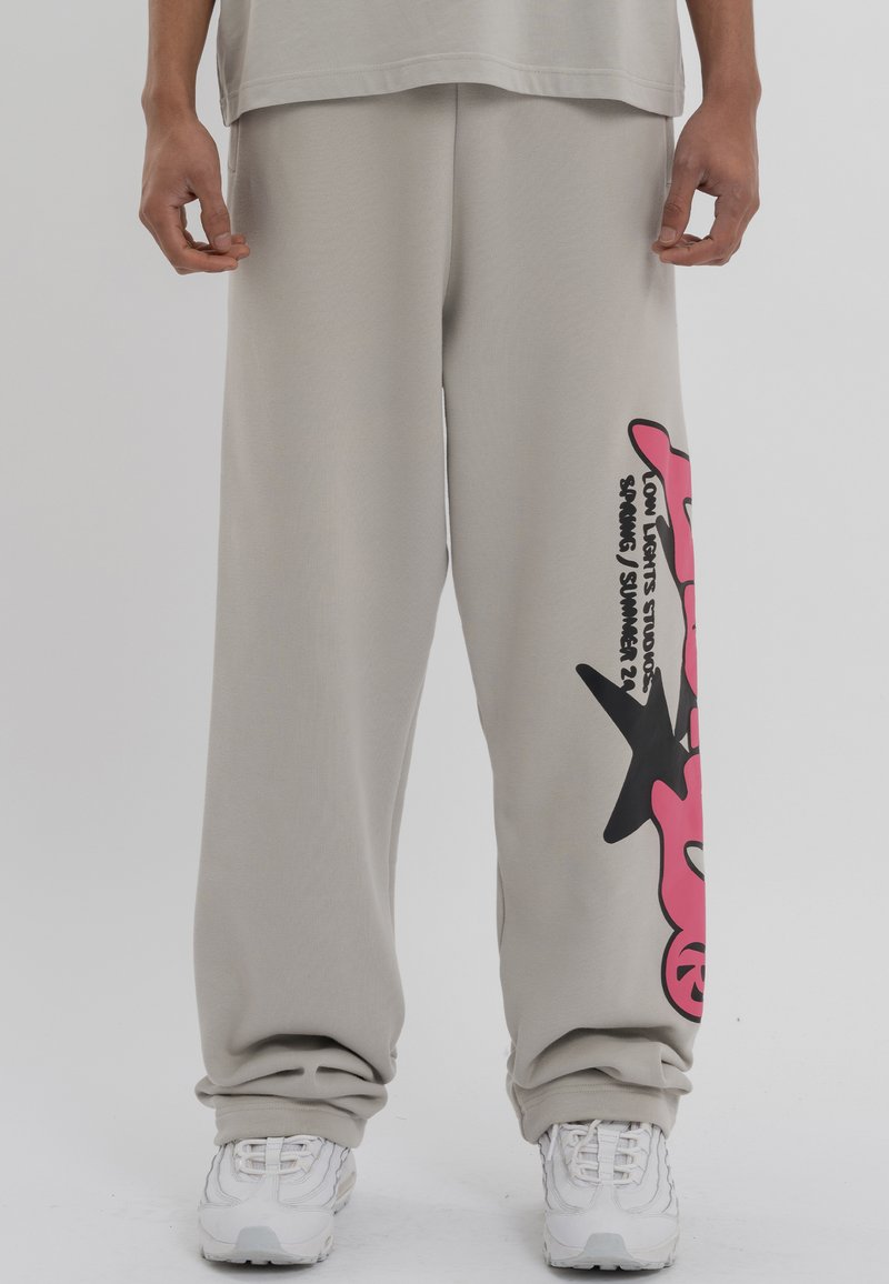 Gaiši pelēkas sporta bikses ar brīvu piegriezumu, uz kreisā kājas ir liela rozā un melna grafika. Uzraksts lasa "LOW LIGHT STUDIOS PAVAIRDI / VASARA 22."