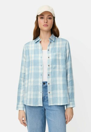 Młoda kobieta w białej czapce, jasnoniebiesko-białej koszuli w kratę z guzikami, białym topie i jasnoniebieskich dżinsach na jednolitym tle.