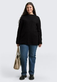 Lachende vrouw met lang donker haar, gekleed in een zwart trui, blauwe jeans en zwarte laarzen, die een beige draagtas vasthoudt tegen een effen lichte achtergrond.