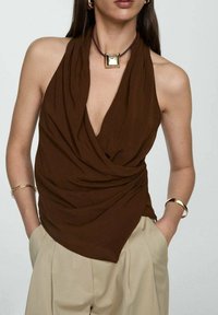 Top a halter marrone con tessuto drappeggiato, caratterizzato da un profondo scollo a V. Abbinato a pantaloni beige a vita alta e accessori in gioielli dorati.