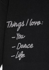 Czarny sweter dziany z białym haftowanym tekstem: "Rzeczy, które kocham: - Ciebie - Taniec - Życie" ułożonym pionowo na materiale.