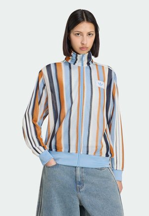 Junge Frau trägt eine Jacke mit Reißverschluss und vertikalen Streifen in Blau, Orange, Schwarz und Weiß, kombiniert mit hellen Jeans.