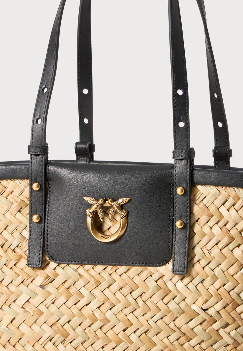 Gewebte Strohtasche mit schwarzen Lederelementen, einem runden goldfarbenen Hardwaredetail und zwei oberen Griffen zum Tragen.