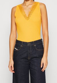 Gelber ärmelloser Body mit einem tiefen V-Ausschnitt, verziert mit texturiertem Spitzenbesatz. Kombiniert mit dunkelblauen, weiten Denim-Jeans.