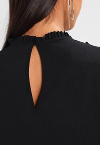 Schwarzes, strukturiertes Oberteil mit gerafftem Ausschnitt und einem Schlitz am Rücken, das glatten Stoff und dezente Details zeigt.
