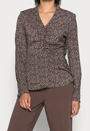 Femme portant un chemisier à manches longues marron à motifs floraux avec un col en V et un pantalon marron, debout devant un arrière-plan uni.