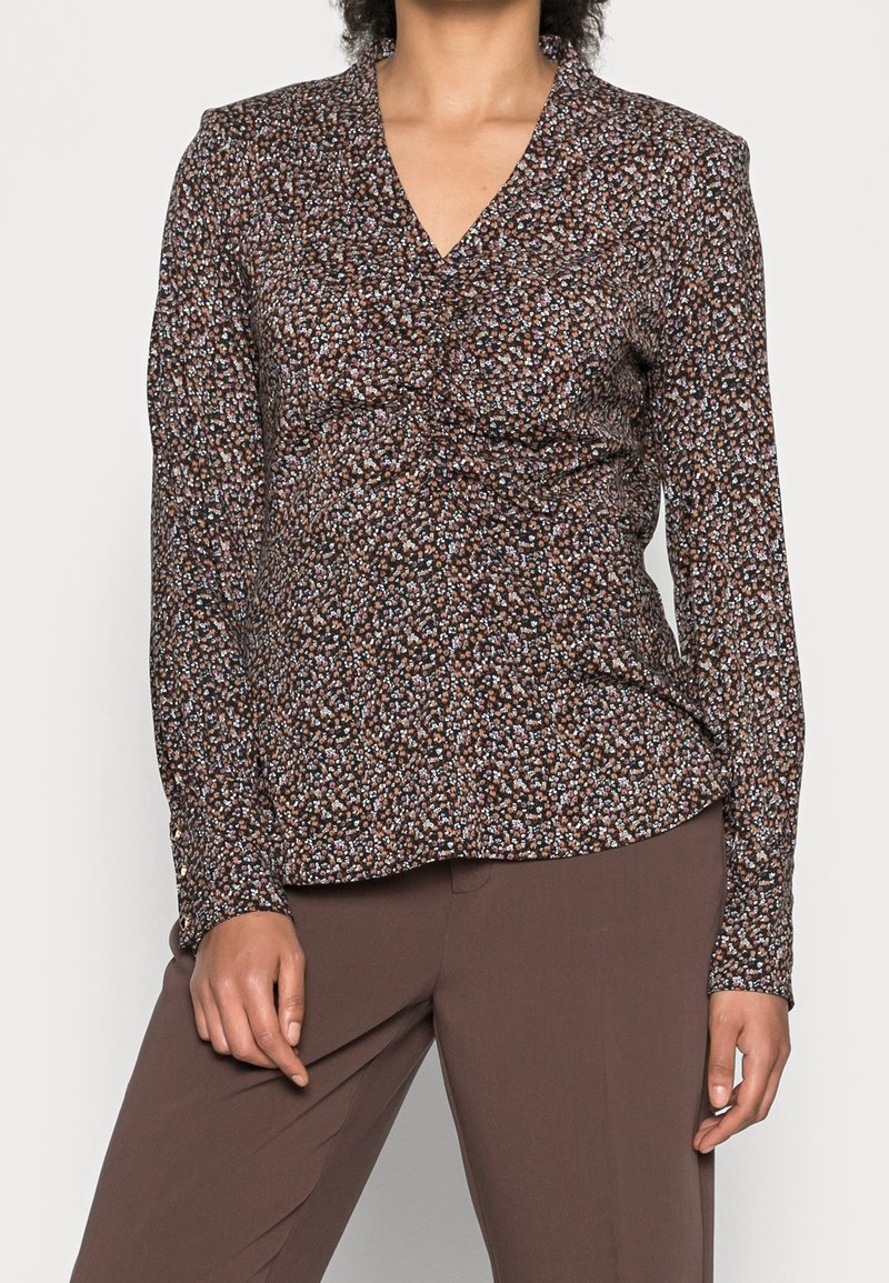 Femme portant un chemisier à manches longues marron à motifs floraux avec un col en V et un pantalon marron, debout devant un arrière-plan uni.