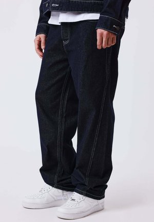 Persona vistiendo jeans holgados de color azul oscuro con costuras blancas en contraste y zapatillas blancas, de pie frente a un fondo liso.