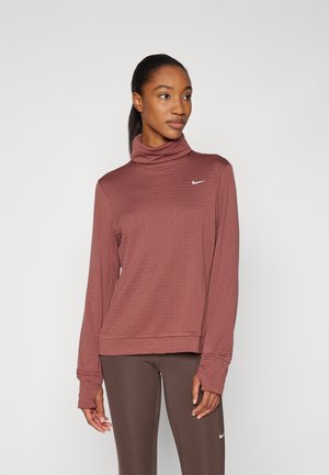 Mujer vistiendo un cuello alto de manga larga color granate y leggings marrones con logos de Nike, de pie contra un fondo claro y liso.