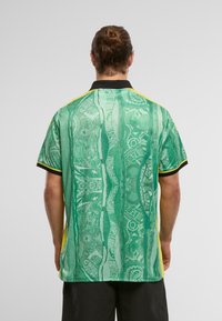 Zöld mintás pólóing fekete gallérral és sárga részletekkel. A design absztrakt formákat és textúrákat tartalmaz, kiemelve egyedi stílusát.