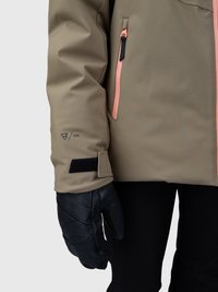 Mâneca de jachetă maro cu fermoar roz și manșetă ajustabilă din Velcro negru, purtată cu mănușă din piele neagră și pantaloni negri, pe un fundal deschis.