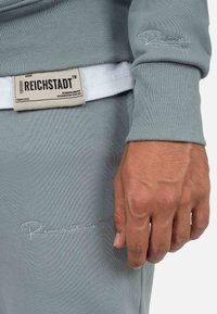 Felpa a coste grigia chiara con dettaglio del logo ricamato. Presenta un'etichetta con "Reichstadt" e istruzioni per la cura, visibile sotto l'abbigliamento.