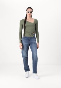 Grön långärmad topp med asymmetrisk halsringning och knapppanel, kombinerad med ljusblå raka jeans och vita sneakers.