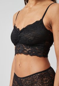 Bralette en dentelle noire avec un design froncé, des bords en forme de vagues et de fines bretelles ajustables. Présente une texture en dentelle transparente et une longueur courte.