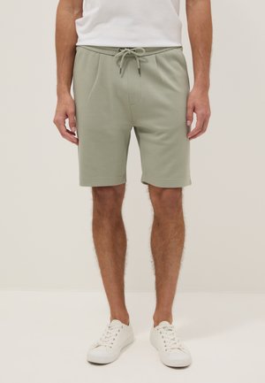 Hombre con pantalones cortos verdes claros con cordón, camiseta blanca y zapatillas blancas, de pie contra un fondo liso y claro.