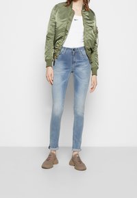 Persona che indossa jeans skinny blu chiaro, sneakers beige, canotta bianca Nike e giacca bomber verde oliva, in piedi contro uno sfondo bianco uniforme.