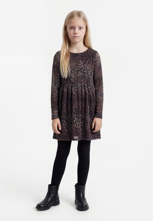 Jauna meitene ar gariem blondiem matiem, valkājot garu piedurkņu leoparda raksta kleitu, melnas zeķbikses un melnus potīšu zābakus, stāvot ar seju pret priekšu.