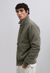 Chaqueta de pana en verde oliva, con cuello alzado, cierre de botones doble y dos bolsillos frontales con solapas.