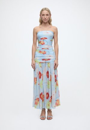 Γυναίκα που φοράει ένα αμάνικο ανοιχτό μπλε φόρεμα με κόκκινο και κίτρινο φλοράλ σχέδιο, στέκεται μπροστά σε ένα απλό λευκό φόντο.