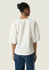 White blouse with short puffy sleeves, textured with square dots. Rounded neckline and a relaxed fit, paired with blue jeans.