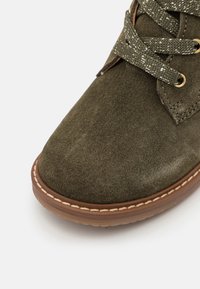 Stivaletti alla caviglia in suede verde con punta rotonda, occhielli in tono oro e lacci testurizzati. Suola cucita color cuoio con impunture bianche in contraste.