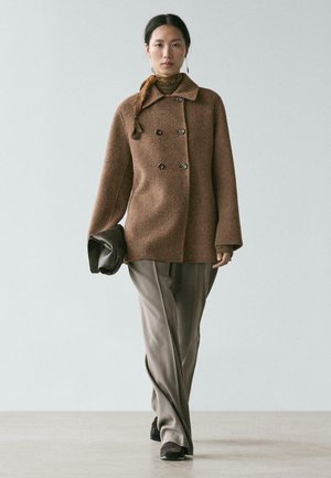 Kvinde går iført en brun dobbeltradet frakke, beige bukser, et tørklæde, holder en sort clutch og har sorte sko på mod en ensfarvet baggrund.