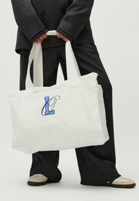 Balta drobės pirkinių kuprinė su mėlynai siuvinėtu "L" logotipu, stačiakampio formos, dviejomis rankenomis ir užtrauktuku viršuje.