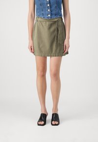 Olivově zelená sukně ke kolenům s překládaným designem a zavazováním na straně. Noste ji s džínovým krátkým topem a černými nazouváky s otevřenou špičkou.