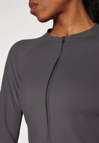 Šedý sportovní top s přiléhavým střihem, dlouhými rukávy a polozipem. Hladká textura podtrhuje minimalistický styl.