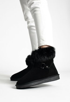 Des bottines en daim noir présentent une bordure en fausse fourrure, une texture lisse et une semelle en caoutchouc plate, avec un subtil accent métallique sur le côté.