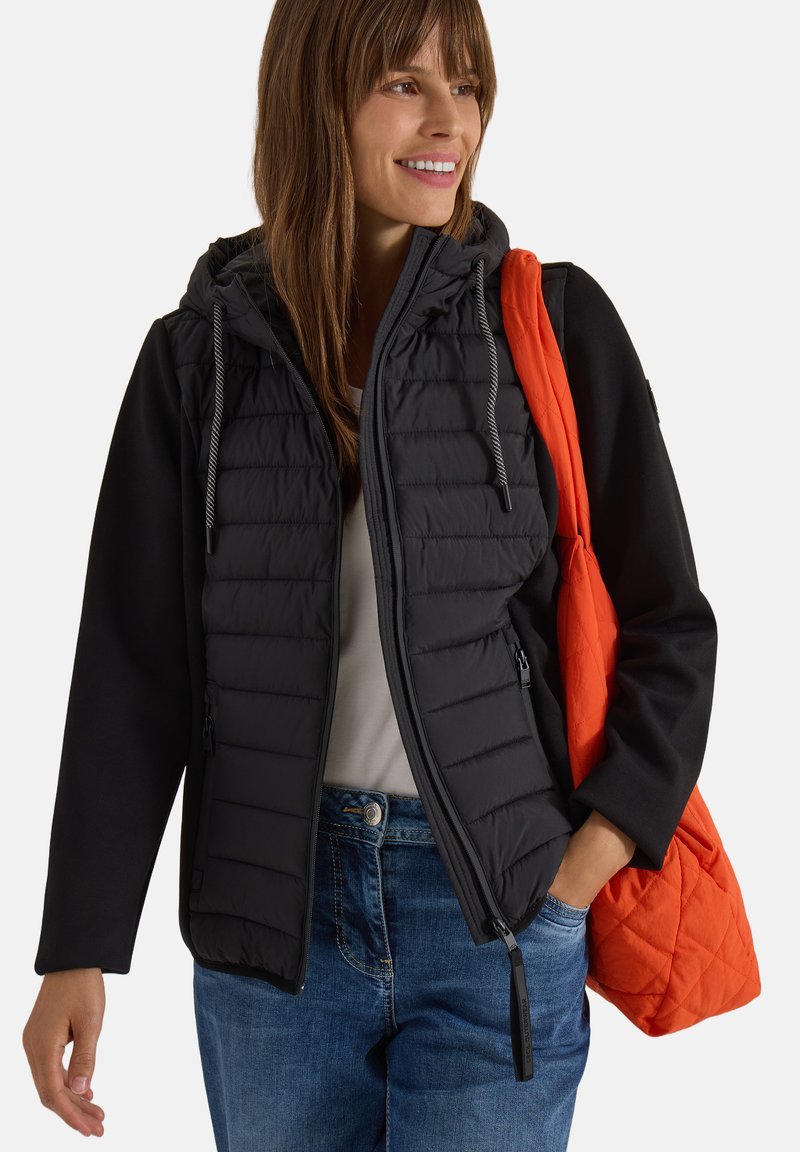 Femme souriante portant une veste matelassée noire avec capuche, un jean bleu, et un sac à épaule matelassé orange sur fond blanc.