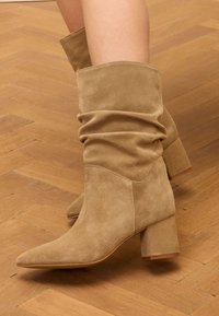 Beige suède enkellaarsjes met een slouchy design, spitse neus en een chunky hak. De textuur is zacht met zichtbare naden en stiksels.