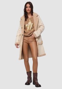 Cappotto in misto lana color crema con colletto in pelliccia, maglione dorato con paillettes e motivo a stelle, pantaloni in ecopelle marroni, e stivaletti alla caviglia marrone scuro.