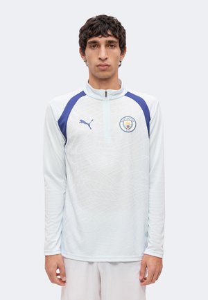 Mand iført en hvid Manchester City træningstrøje med blå Puma-logo og skulderstriber, stående mod en ensfarvet baggrund.