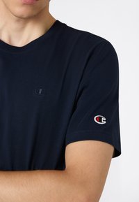 Camiseta de algodón azul marino con cuello en V, que presenta un pequeño logo bordado en el pecho y un logo de color en la manga izquierda.