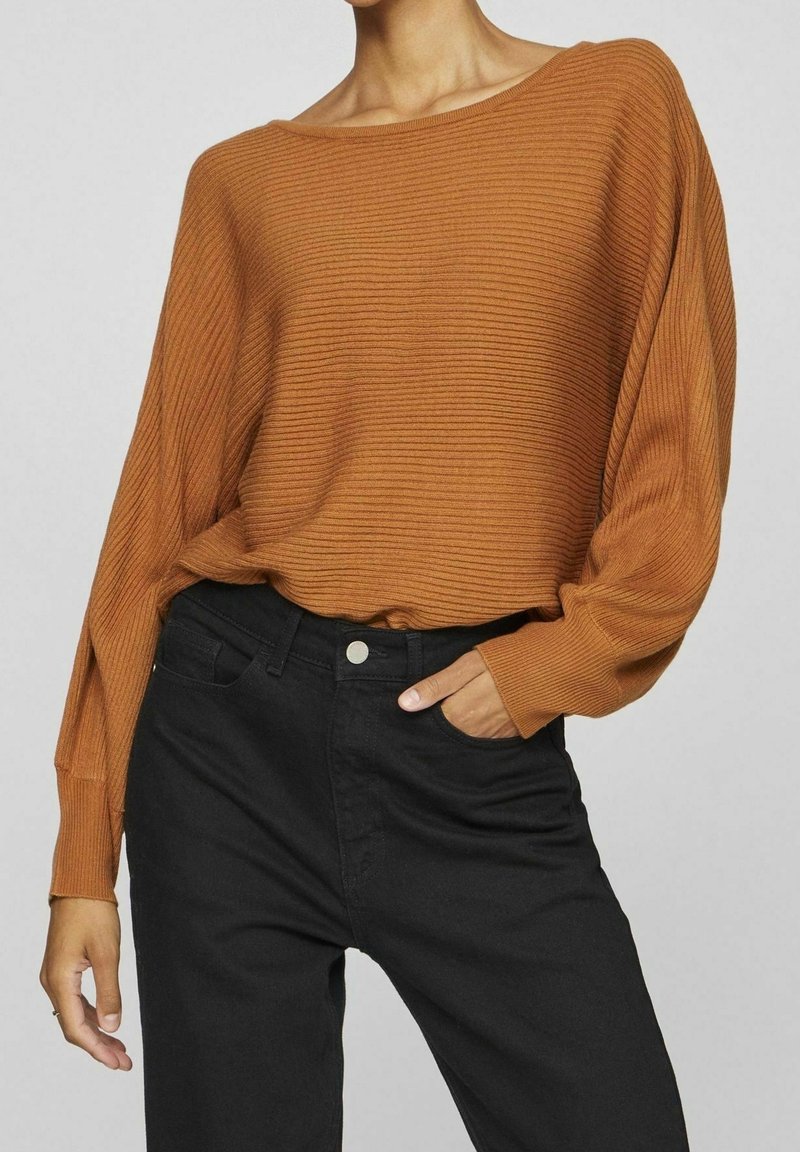 Femme portant un pull côtelé, ample, à manches longues couleur orange brûlé, rentré dans un pantalon noir taille haute, main dans la poche.