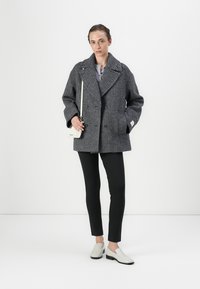 Dobbeltbrystet frakke i grå houndstooth-mønster med brede revers, båret over en lys top og sorte tætsiddende bukser, kombineret med lyse slip-on sko.